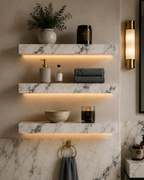 A styled bathroom interior with three floating marble shelves mounted on a beige textured wall, each shelf illuminated by warm under-shelf lighting.