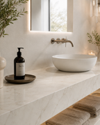 A modern bathroom vanity setup with a white oval vessel sink on a light marble countertop, a dark amber soap dispenser on a tray, folded towels, and warm ambient lighting.